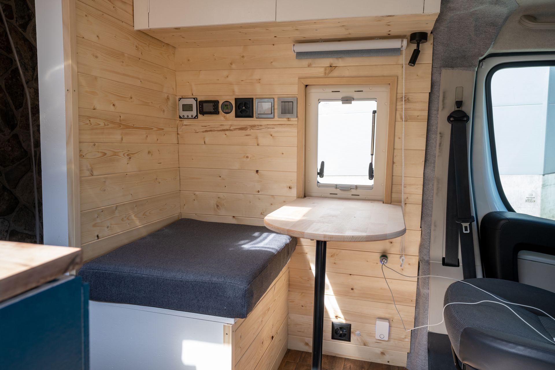 Compact wooden camper van interior with bench bed, small table, and window seat