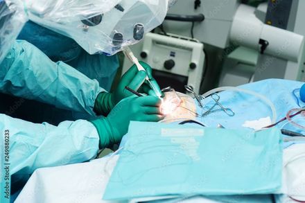 Cirurgiões com luvas verdes e aventais azuis realizam uma operação sob luzes brilhantes da sala de cirurgia.