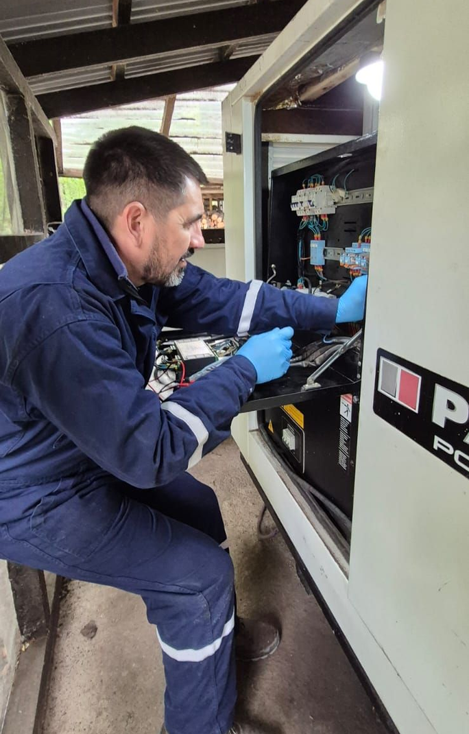 Técnico con mono azul y guantes inspeccionando equipos dentro de un armario eléctrico abierto.