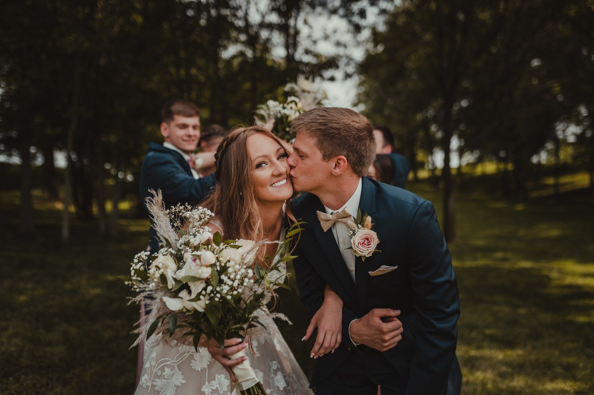 Newly married couple smiling, groom kissing bride's cheek, holding bouquet, forest background.