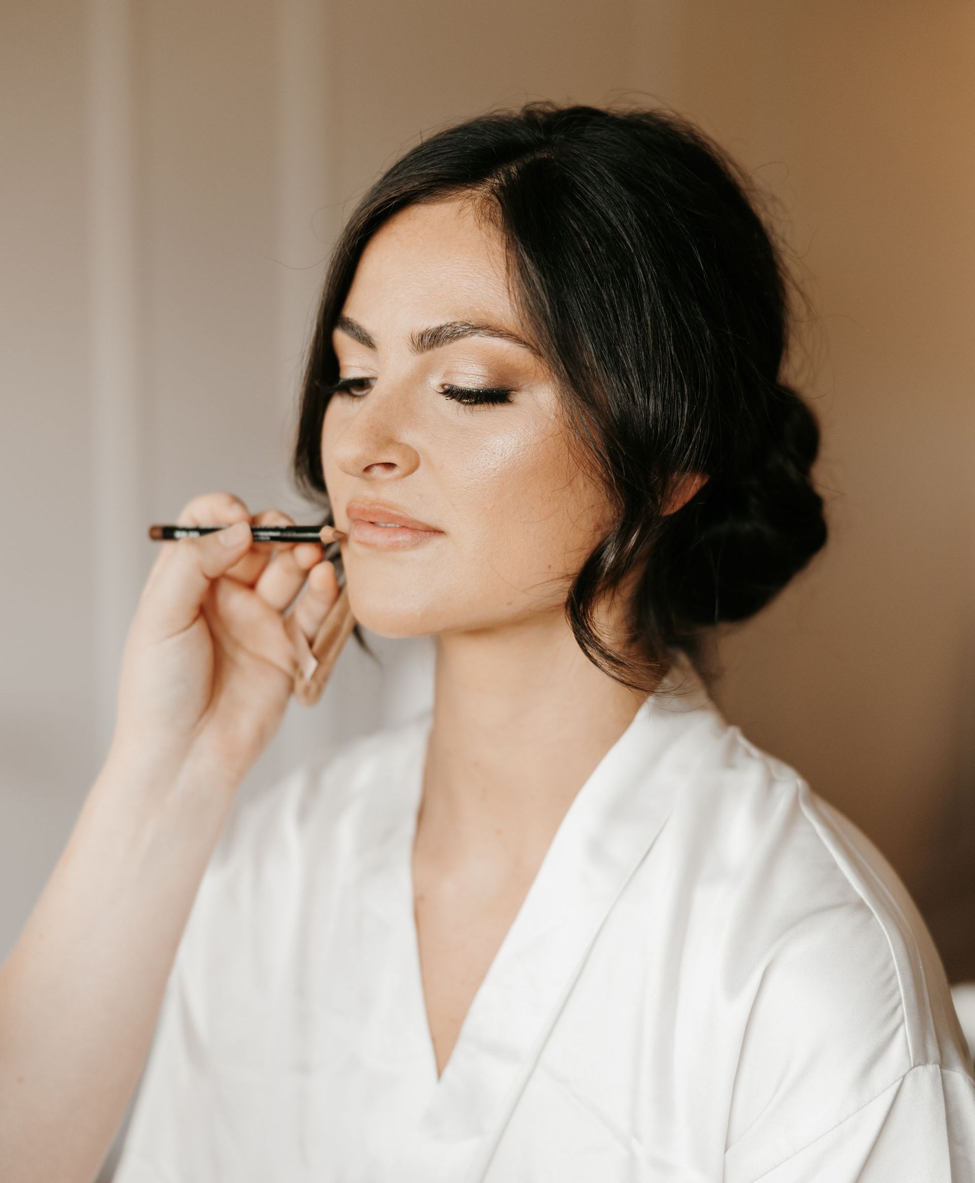 Woman in white robe having makeup applied; neutral expression, indoor setting.