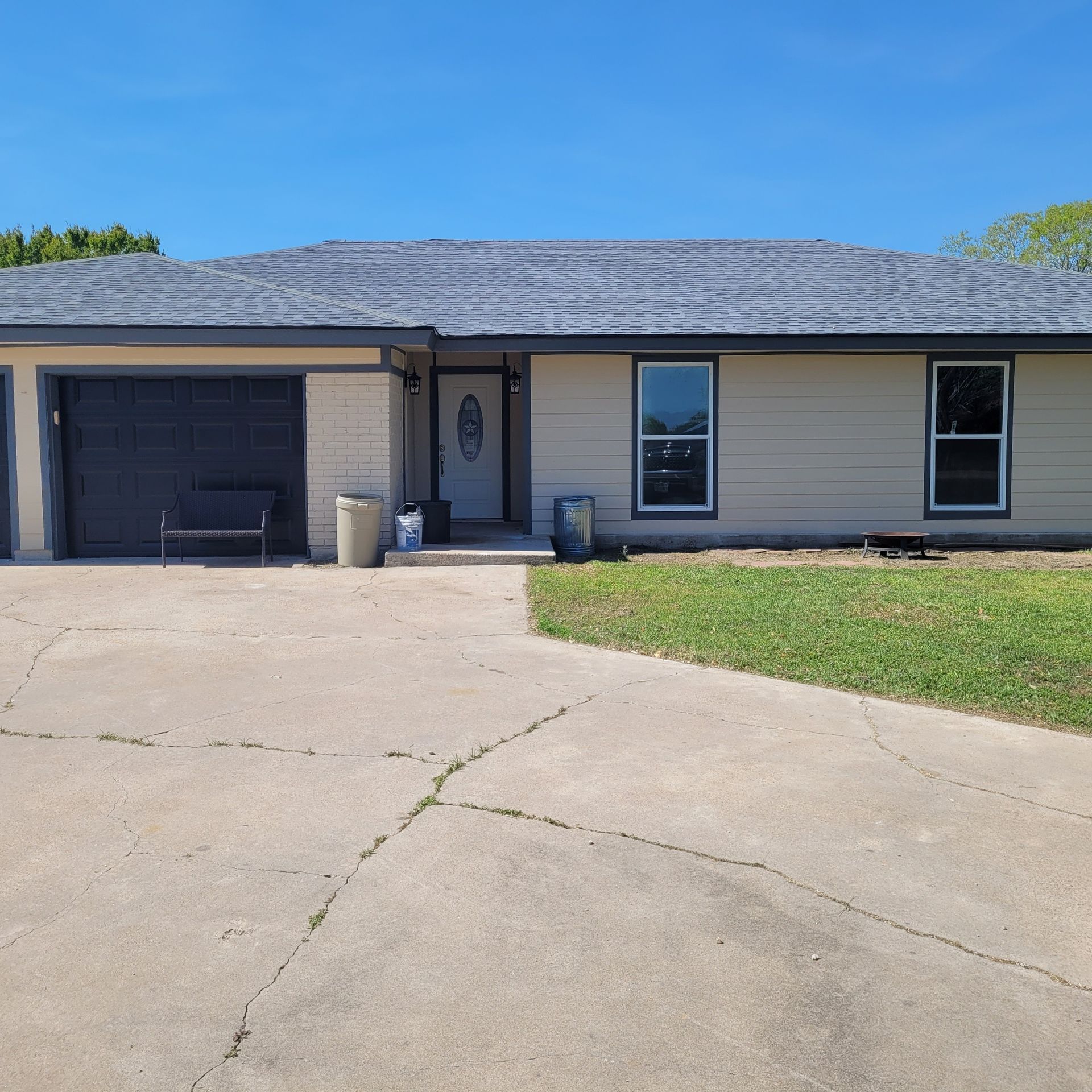 Single-story beige house with a dark gray garage door, driveway, and small front lawn under a blue sky