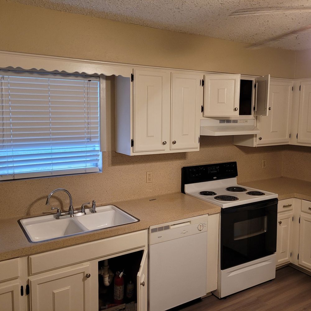 Small beige kitchen with white cabinets, sink, dishwasher, and white stove under a window with blinds.