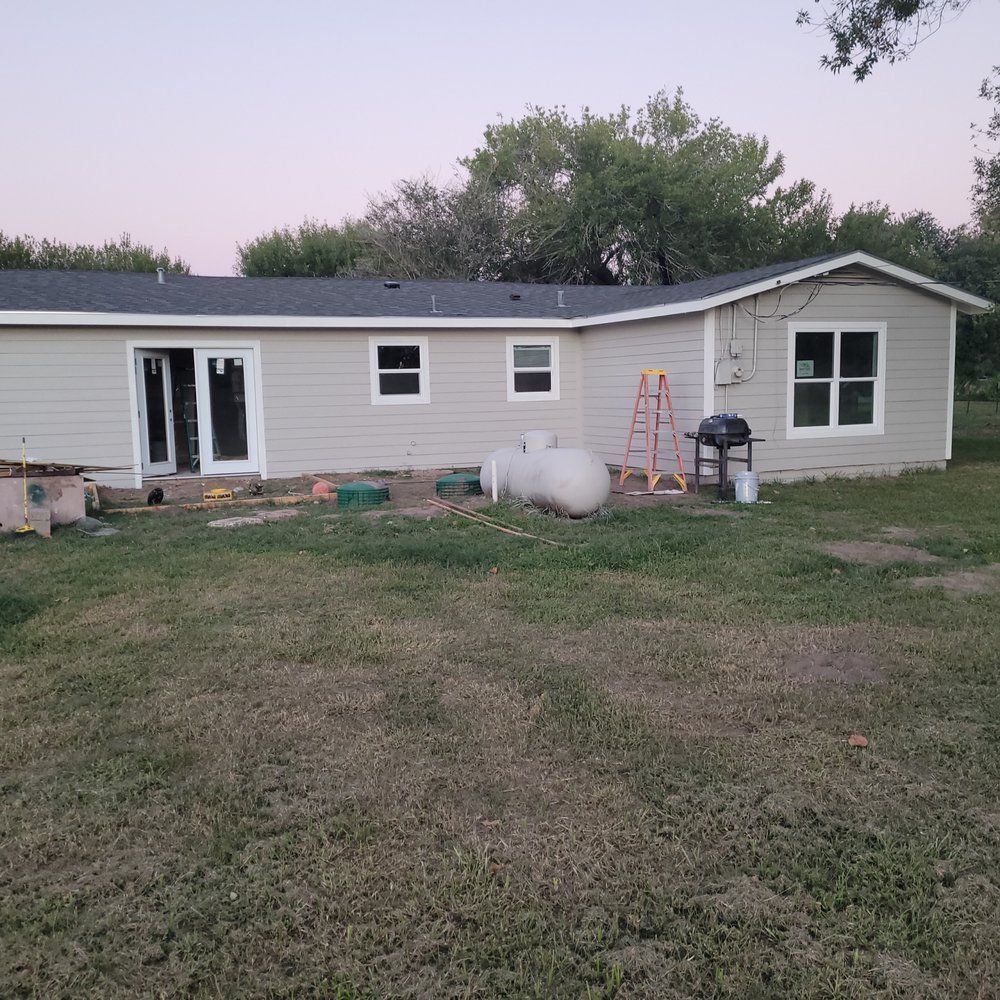 Single-story beige house with white trim, a ladder, grill, propane tank, and an unfinished yard at dusk.