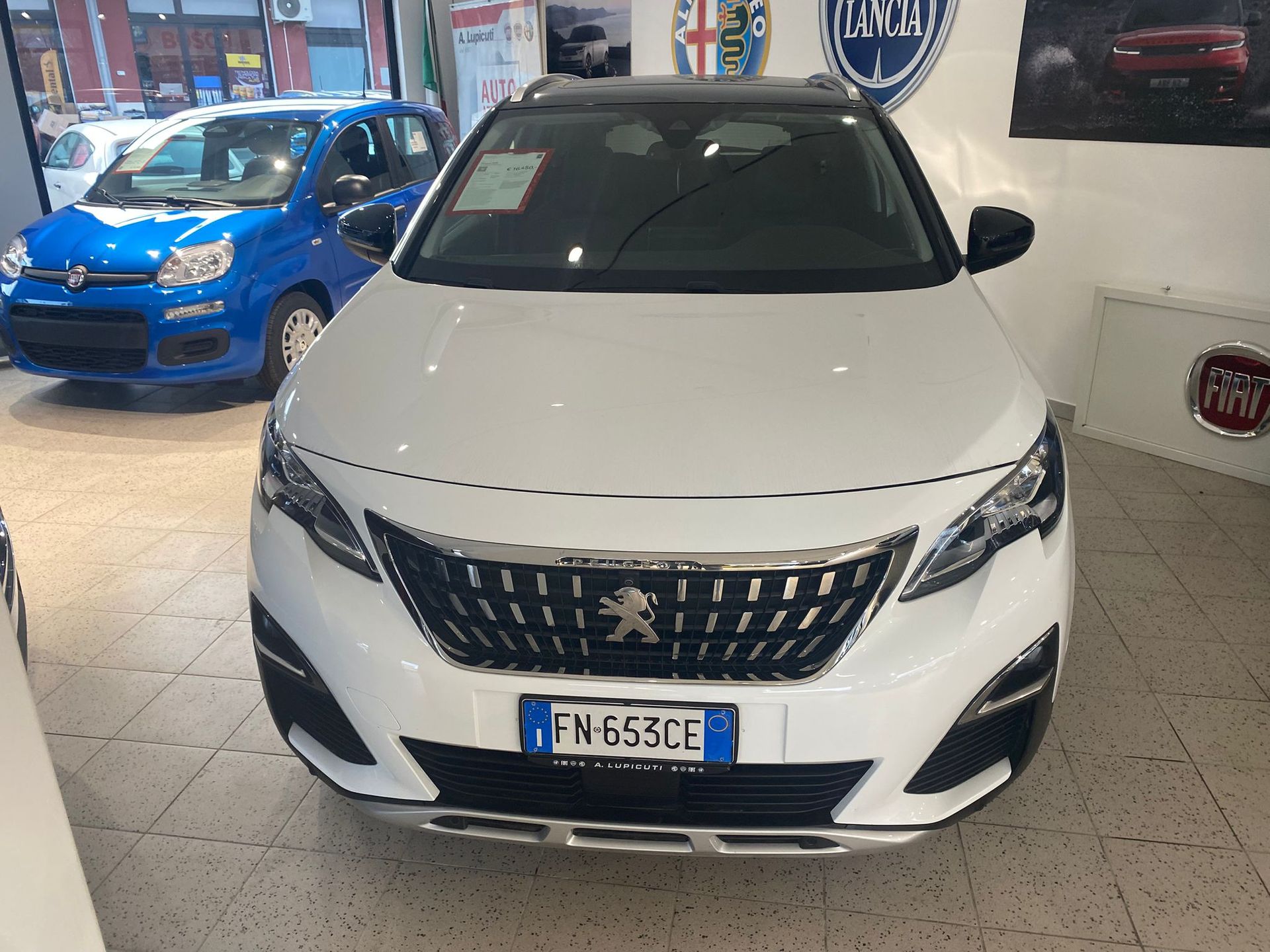 Berlina Peugeot bianca esposta in uno showroom, vista frontale, con un'auto blu sullo sfondo.