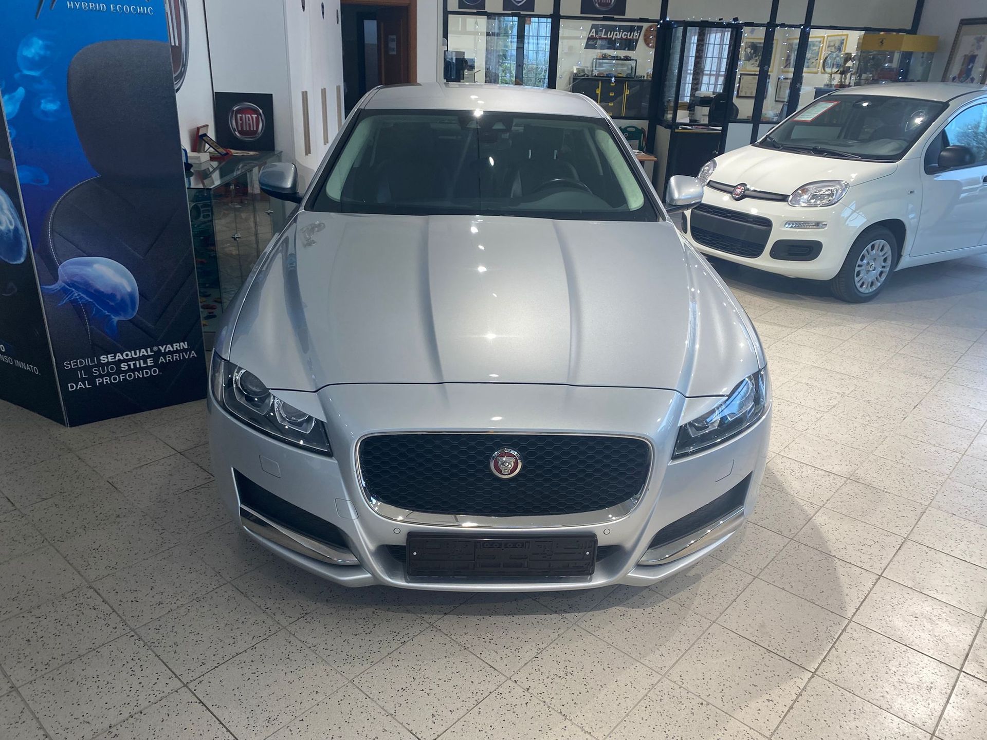 Berlina Jaguar color argento esposta in una concessionaria, vista frontale su un pavimento piastrellato.