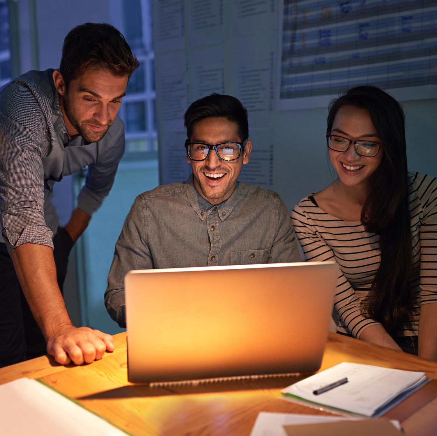 Three coworkers gathered around a laptop, smiling in a dim office lit by the screen.