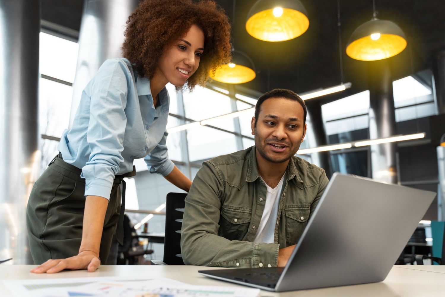 Two coworkers reviewing a laptop in a modern office with hanging yellow lights