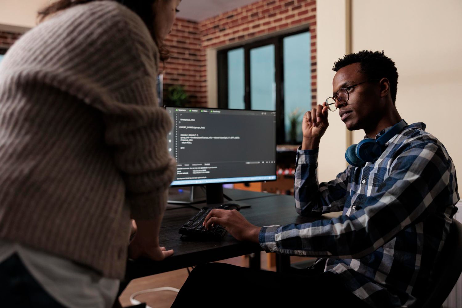 Two people work at a desk with a computer in a brick-walled office, one reviewing code on screen.
