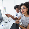Two coworkers in a bright office pointing at a desktop monitor during a discussion