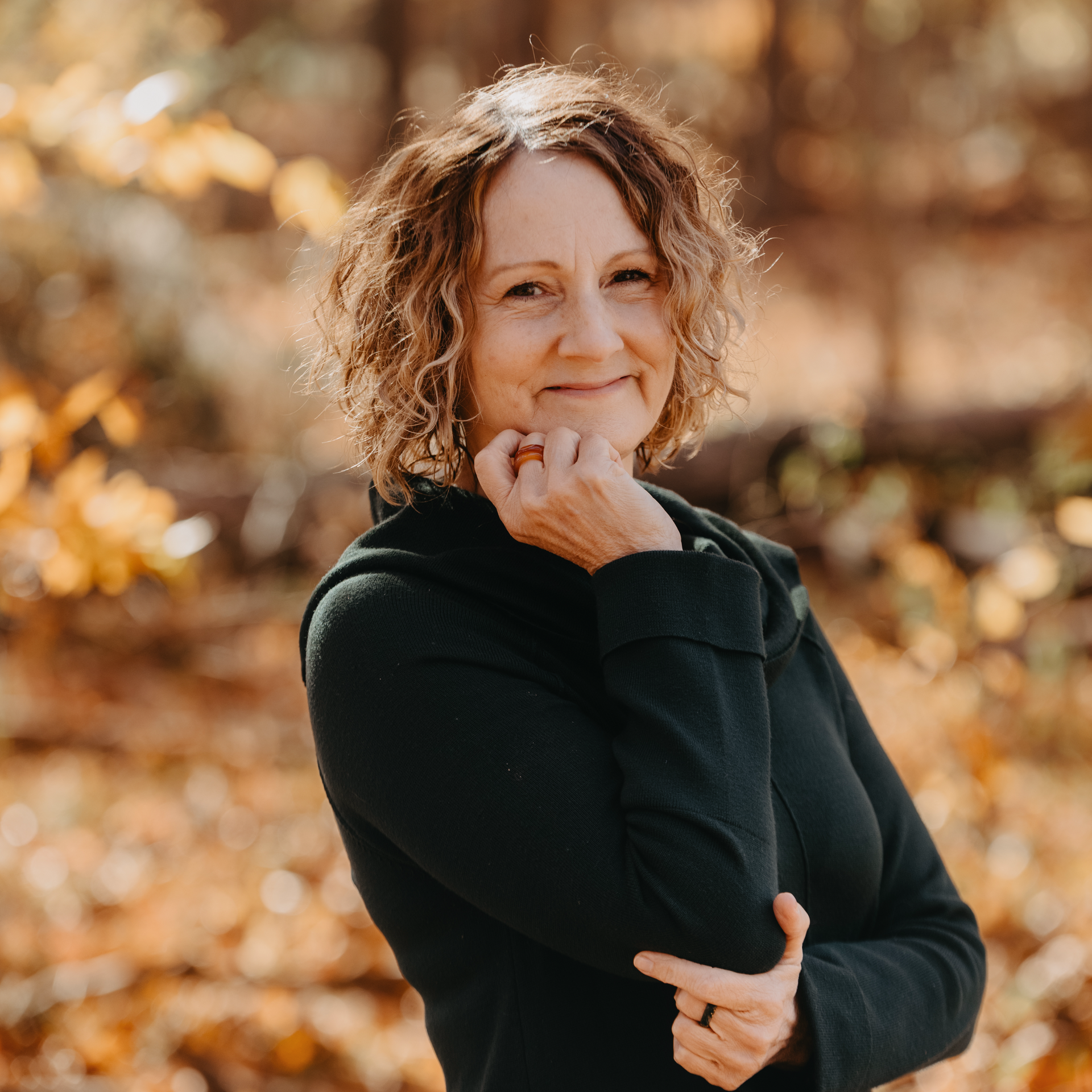 Woman with curly brown hair, smiling, resting chin on hand, wearing a dark green top, outdoors in autumn.