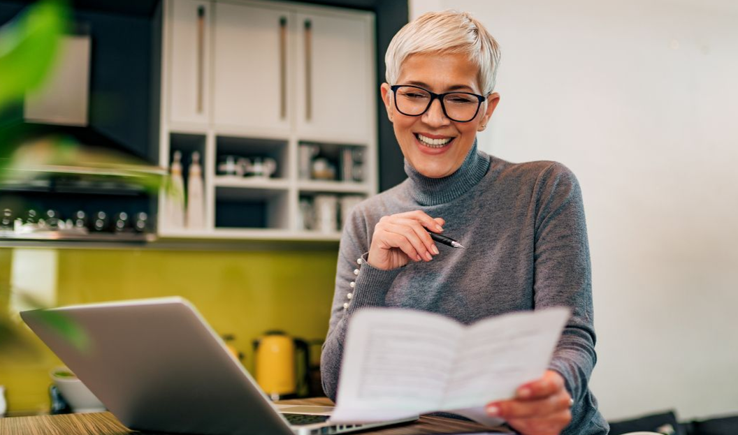 Woman with glasses smiling at paperwork, laptop open on counter, kitchen background.