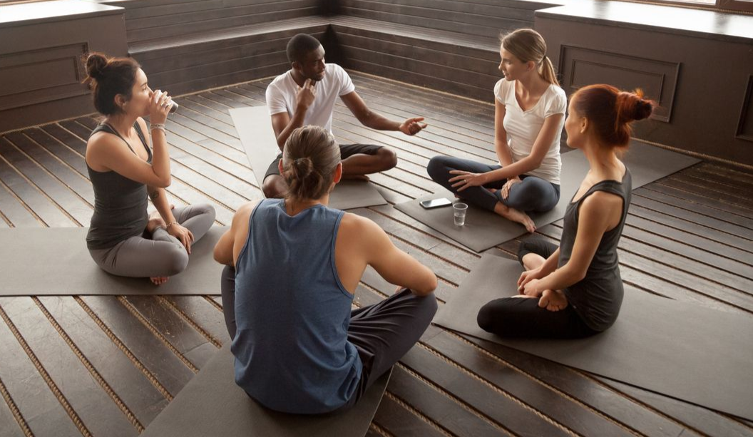 Group of people sitting in a circle on yoga mats, talking and gesturing in a studio.