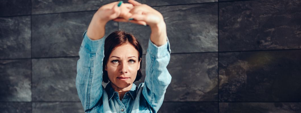 Woman in a blue denim shirt stretches arms overhead, against a gray tiled wall.