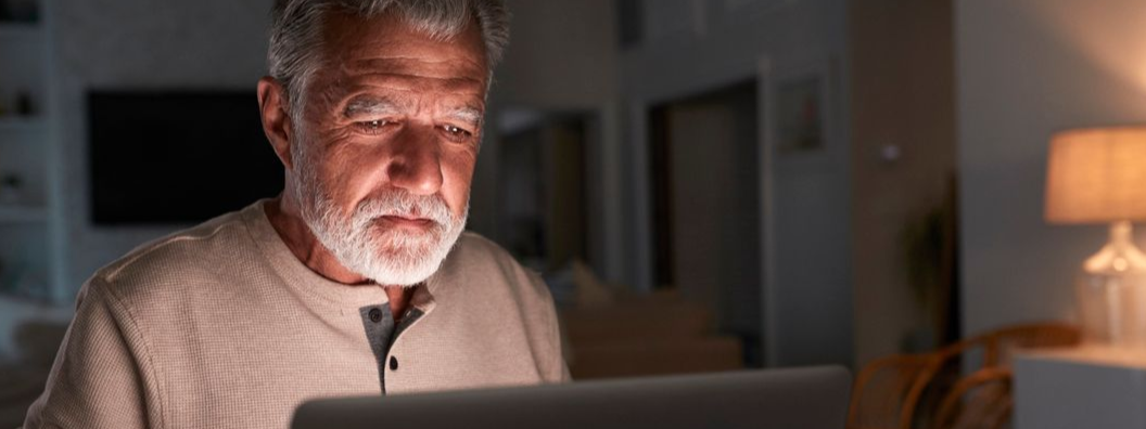 An older person looks at a laptop screen in a dim room, illuminated by the screen and a nearby lamp.