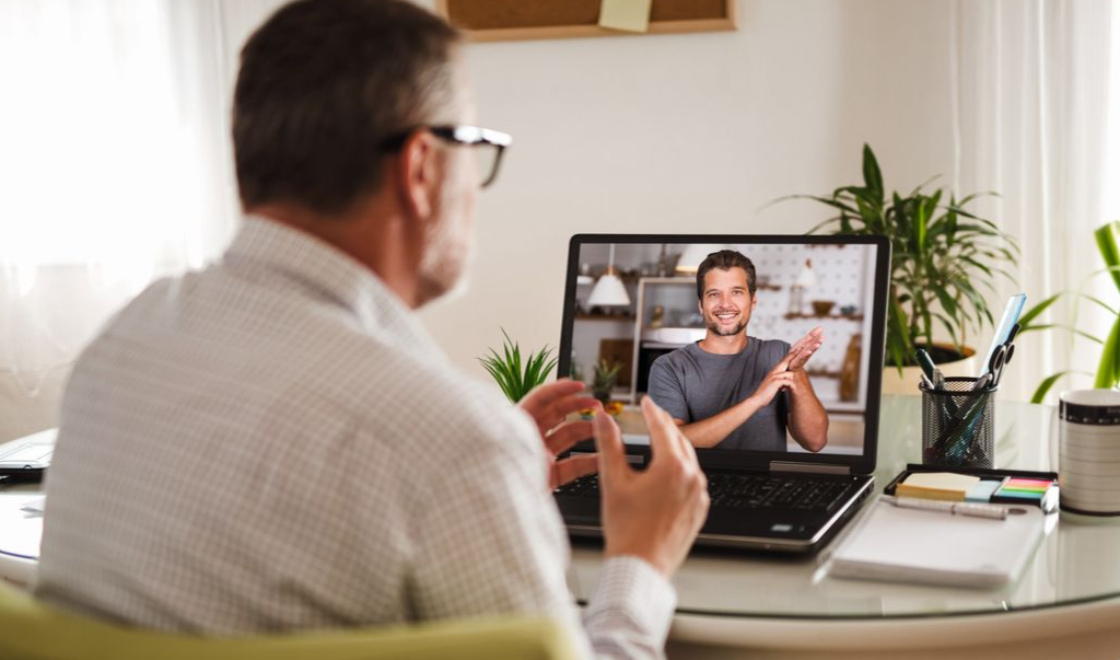 Man in glasses on video call with another man, who is clapping. Laptop on desk, plants nearby.