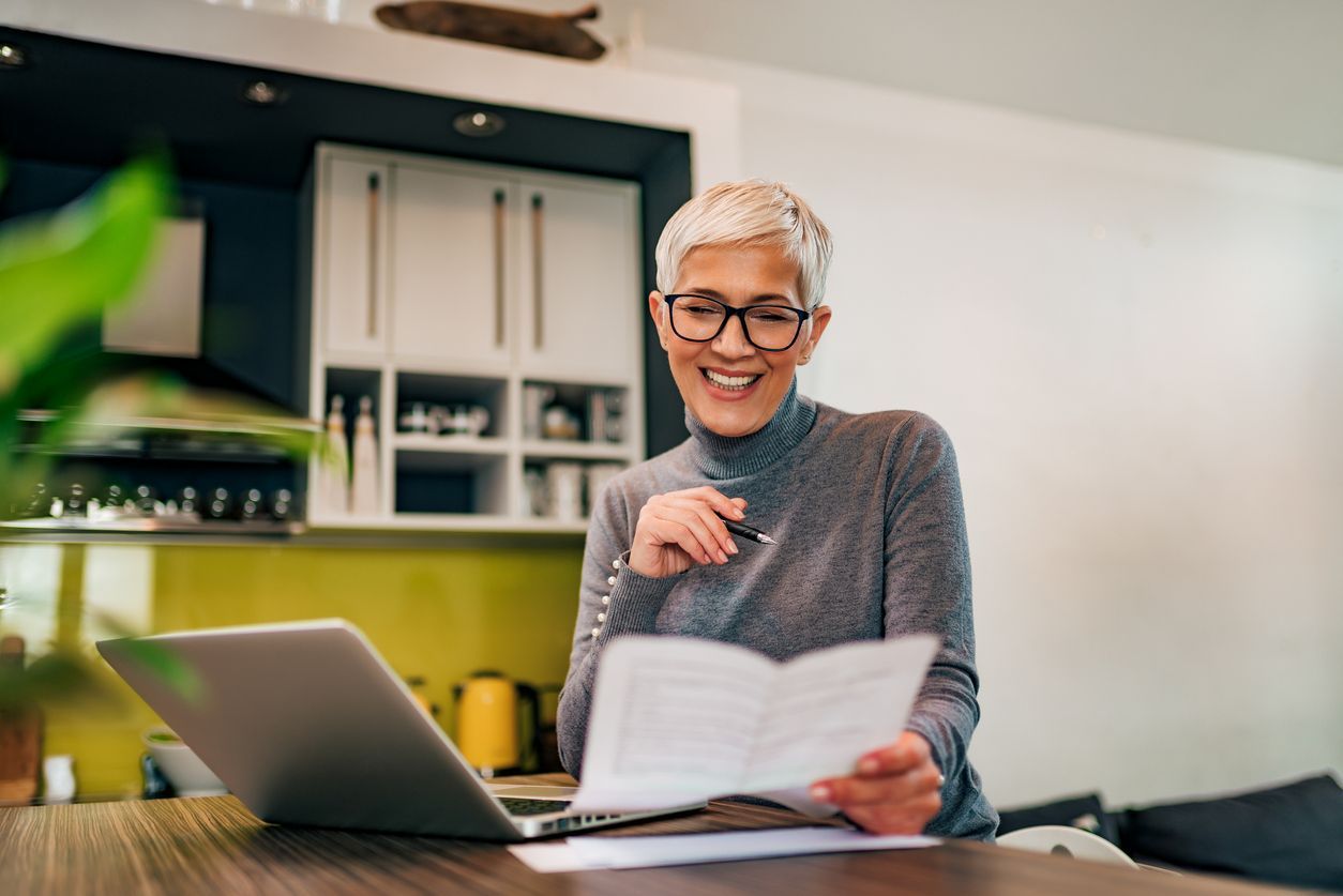 Woman with glasses smiles while reviewing documents near a laptop and kitchen.