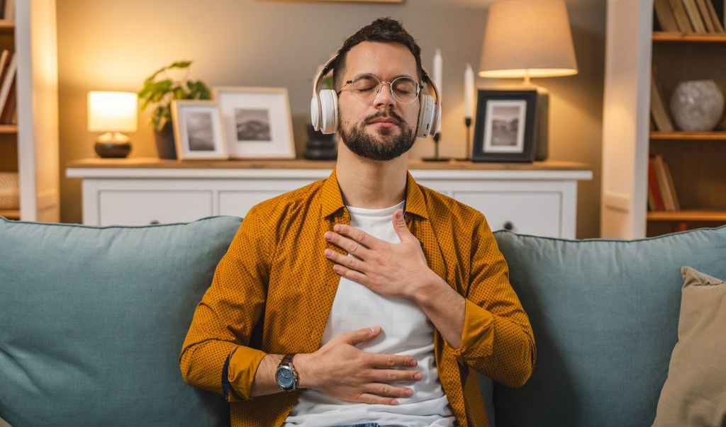 Man wearing headphones, meditating on a sofa, eyes closed, hands on chest and abdomen.