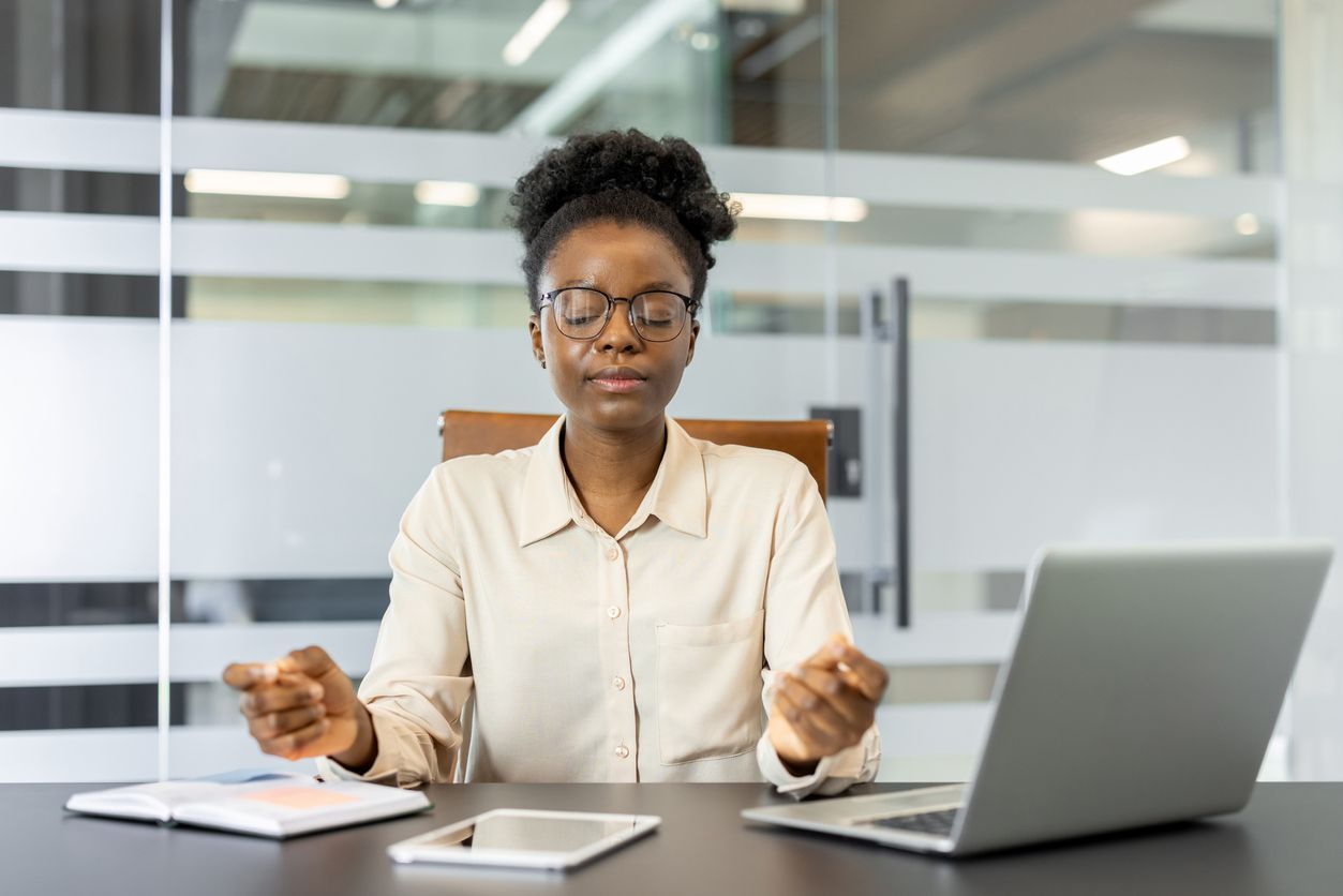 Woman meditating at a desk, eyes closed, hands in fists, laptop and tablet present, office setting.