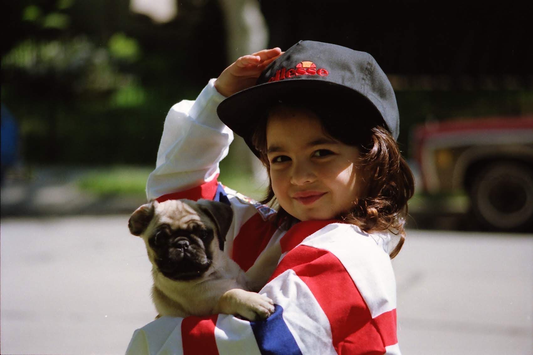 Child in red-and-white shirt holding a pug and adjusting a black cap outdoors