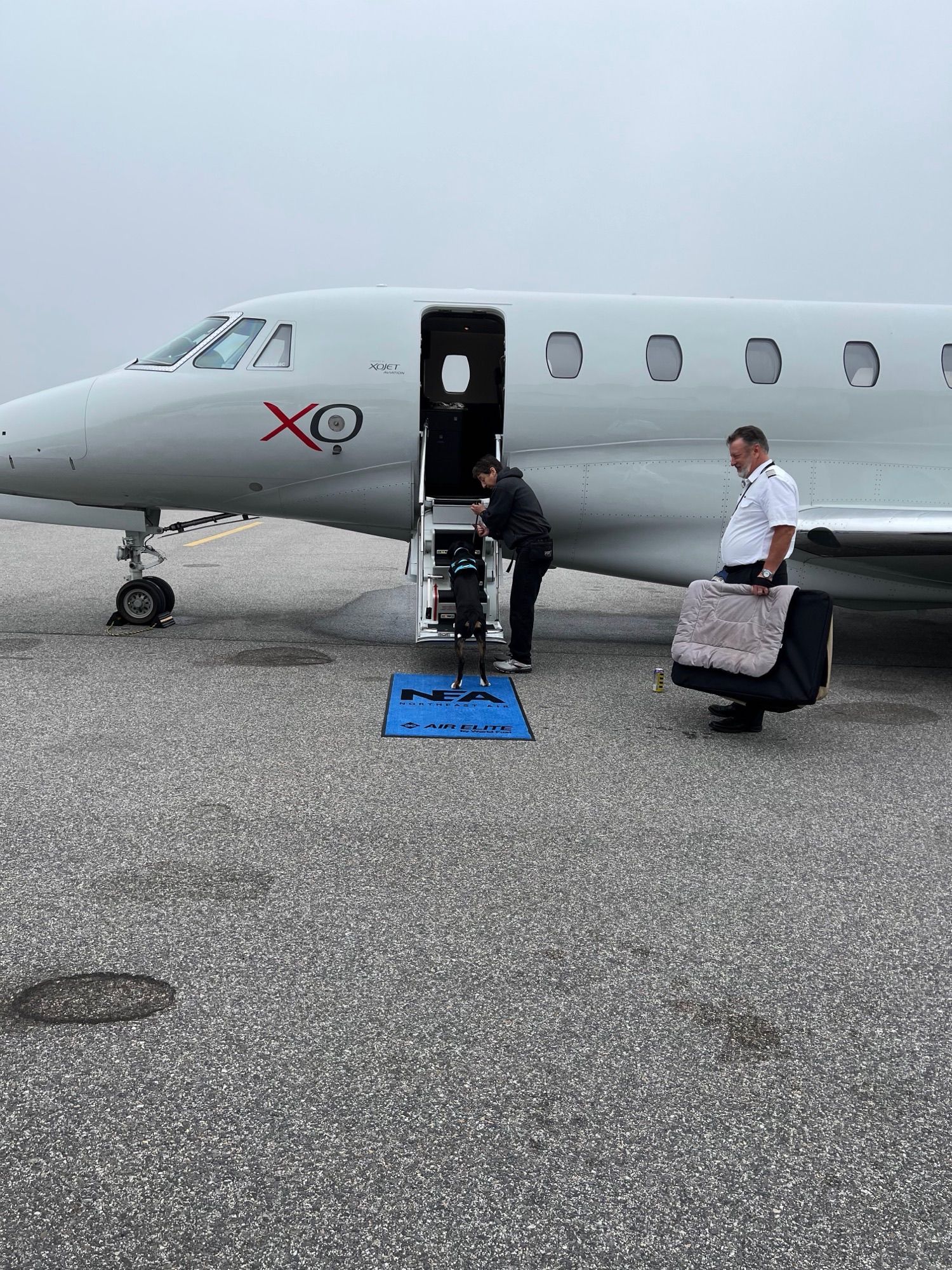 Small jet on tarmac with open door and stairs; a person stands nearby holding a bag.