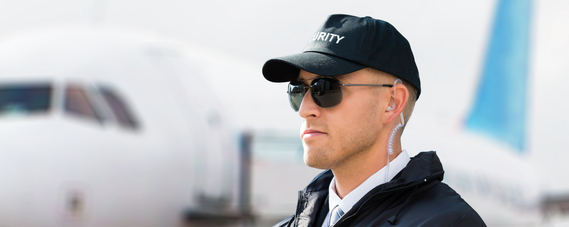 Man in black cap and sunglasses at an airport, with a plane blurred in the background