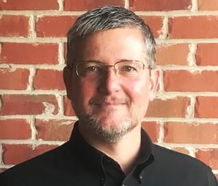 Portrait of a smiling man with glasses in a black shirt against a red brick wall.