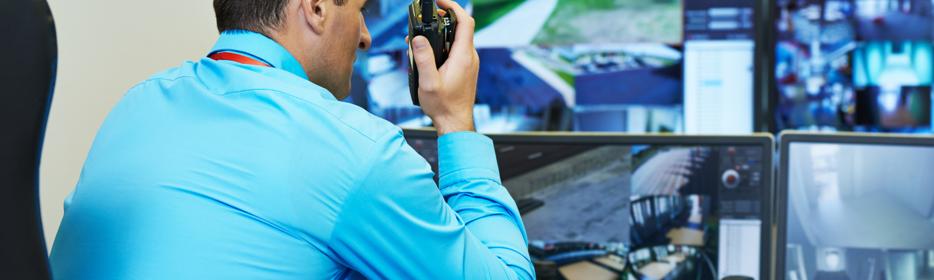 Man in a blue shirt holding a camera near monitors, viewing surveillance footage in a control room