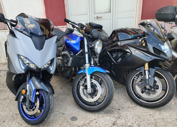 Tre motociclette parcheggiate una accanto all'altra davanti a un edificio: uno scooter argentato, una moto blu e una moto nera.