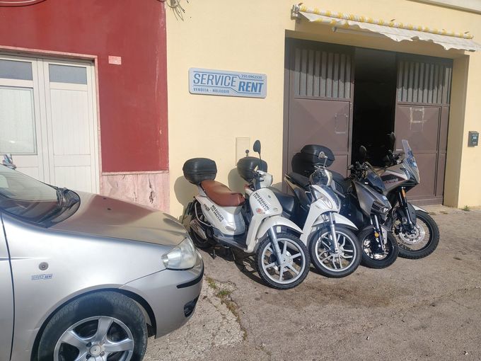 Diversi scooter parcheggiati e un'auto argentata davanti a un edificio rosso con un garage aperto.