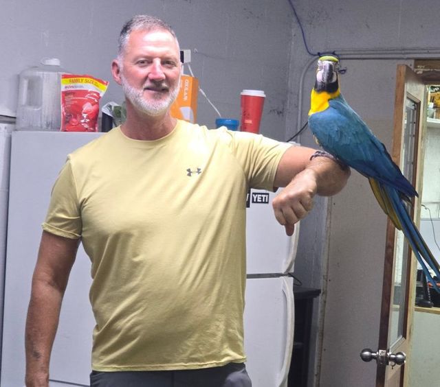 Man standing indoors with a blue-and-yellow parrot perched on his arm | Pine Island Auto