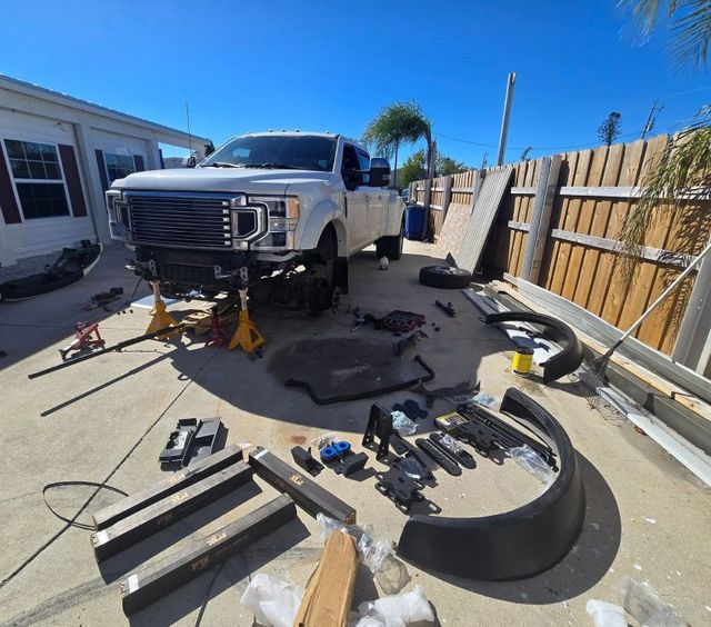 Truck repair parts scattered on driveway beside a white pickup and house, with fence and tools in sunlight. | Pine Island Auto