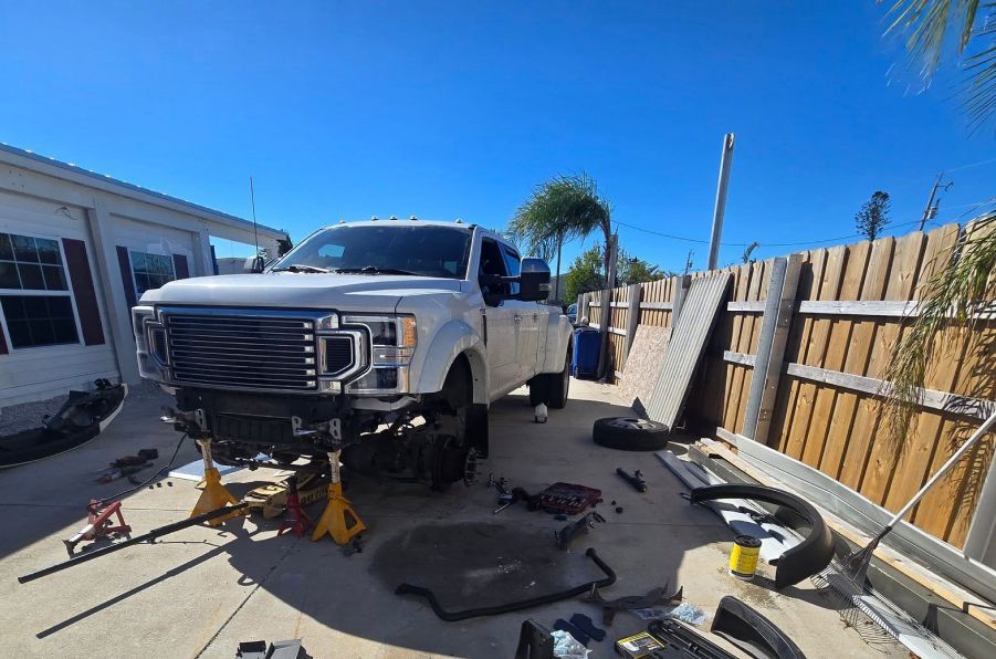 White pickup truck on jack stands in a driveway, surrounded by auto parts and tools near a fence. | Pine Island Auto