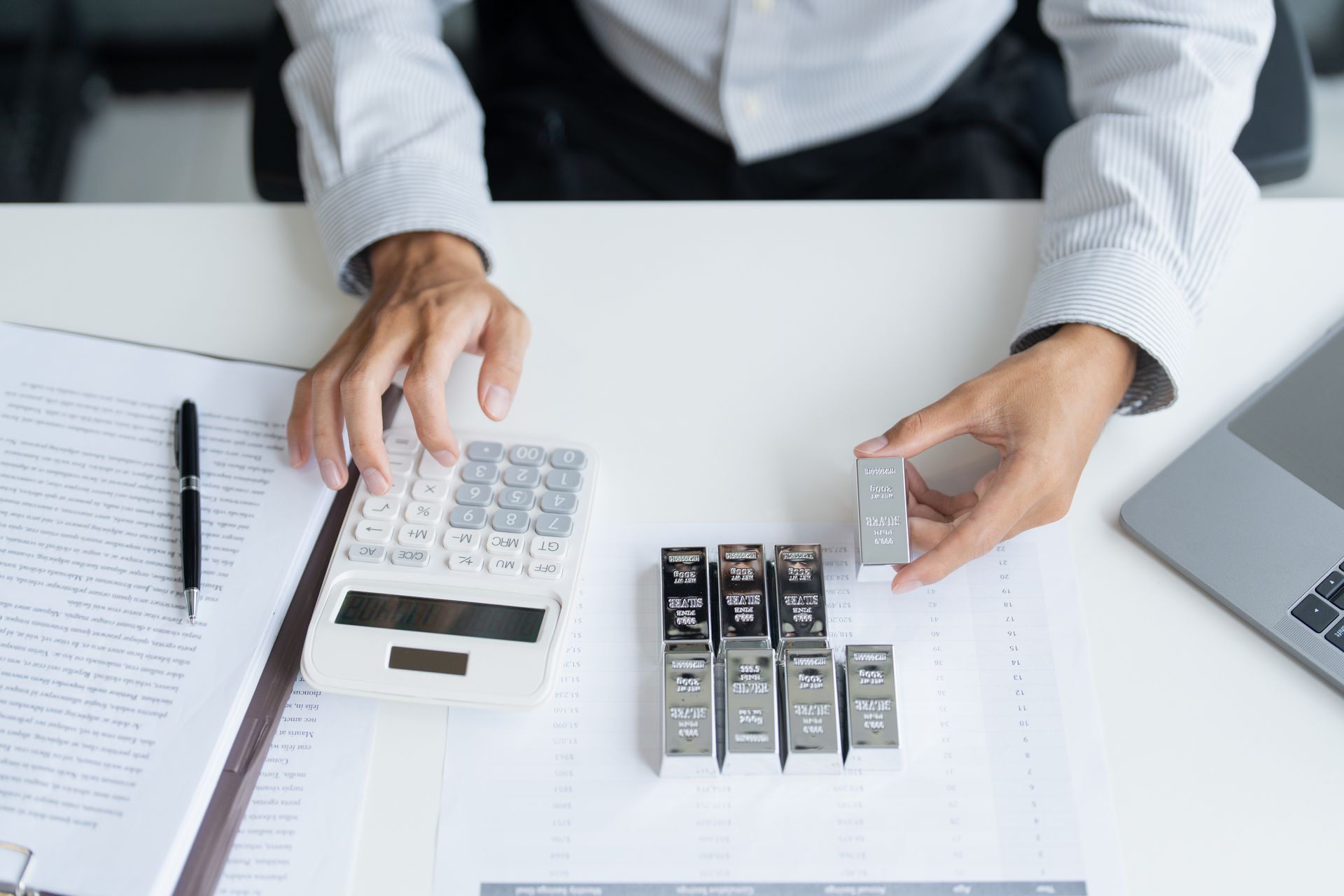 Hands using a calculator to evaluate stacked silver bullion bars on a desk