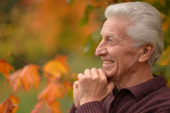 An elderly man is smiling with his hands folded in front of a tree.