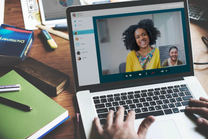 A laptop showing a video call with two people on a wooden desk with a notebook, pen, and books.