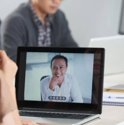 A person viewing a video call on a laptop; the screen shows a smiling man in a white shirt resting his chin on his hand.