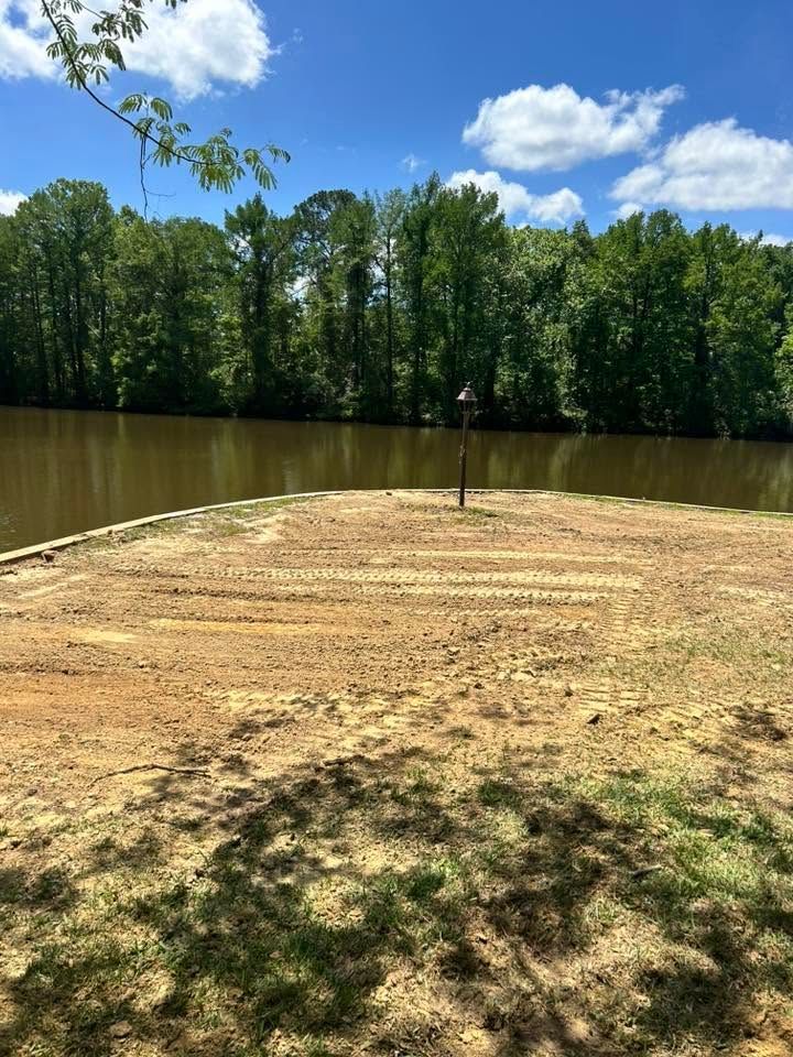 Dirt clearing beside a calm brown river, with a green tree line and blue sky with clouds beyond.