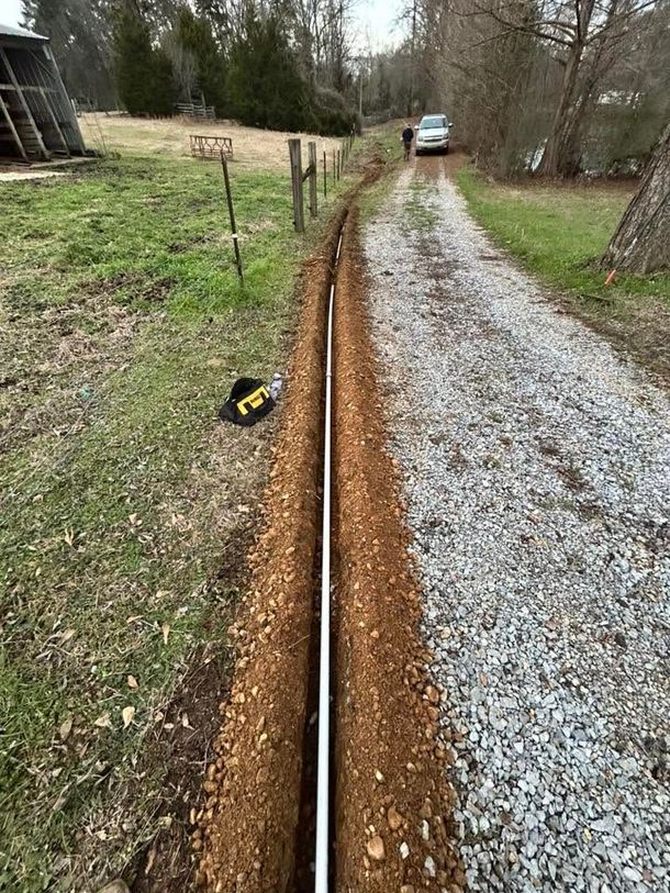 Long gravel driveway with a shallow trench along the left side, a parked car in the distance, and trees nearby.