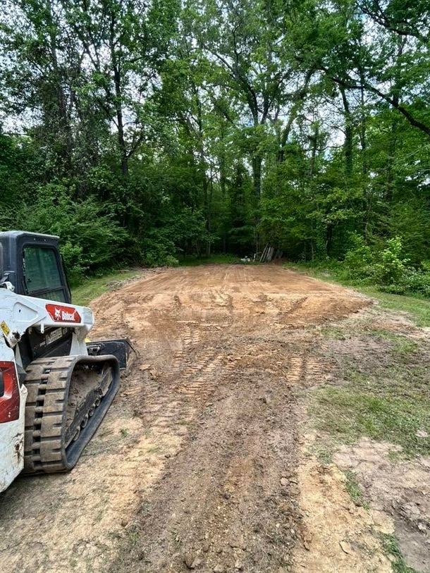 A tracked skid steer at left near a dirt clearing surrounded by dense green trees.