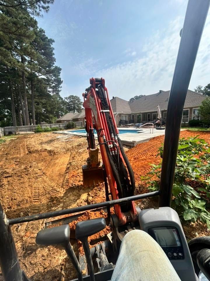 Excavator digging red clay beside houses and trees on a construction site