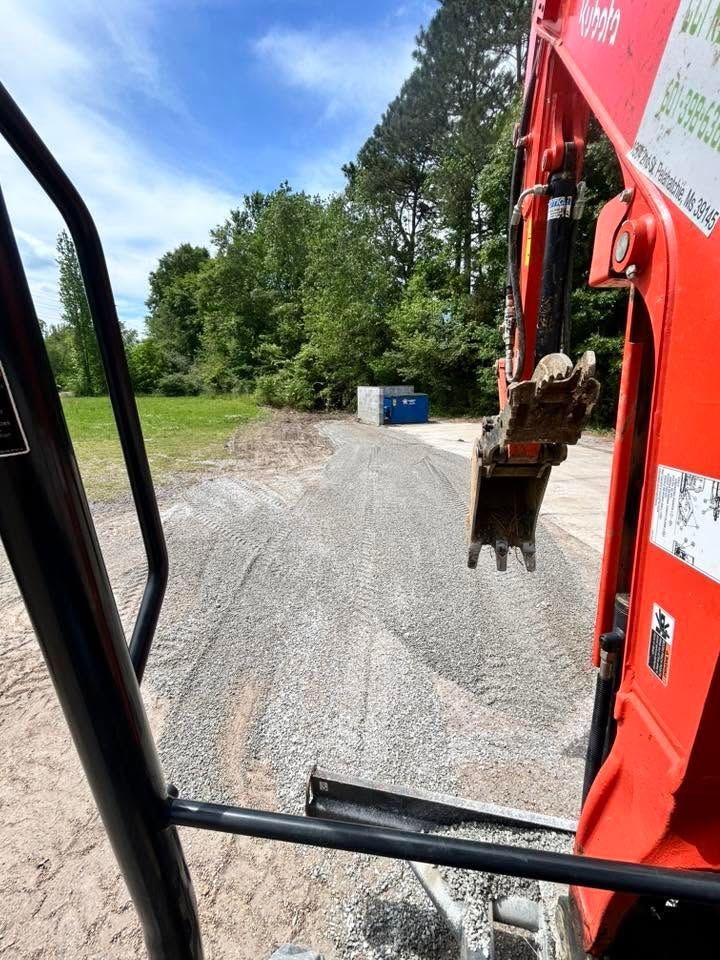 View from a red excavator over a gravel path toward trees and a blue dumpster under a bright sky