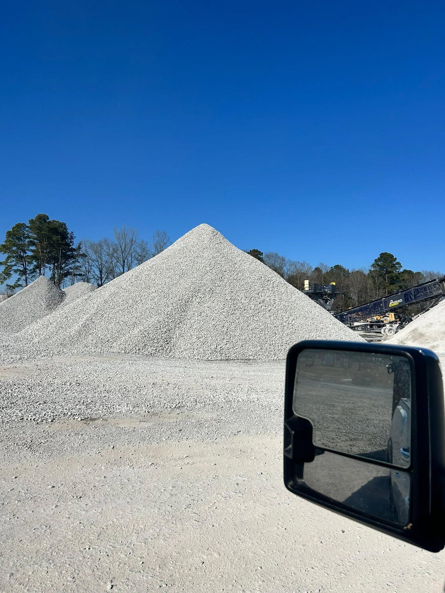 Large white gravel pile in a sunny outdoor lot, seen beside a vehicle mirror under a blue sky