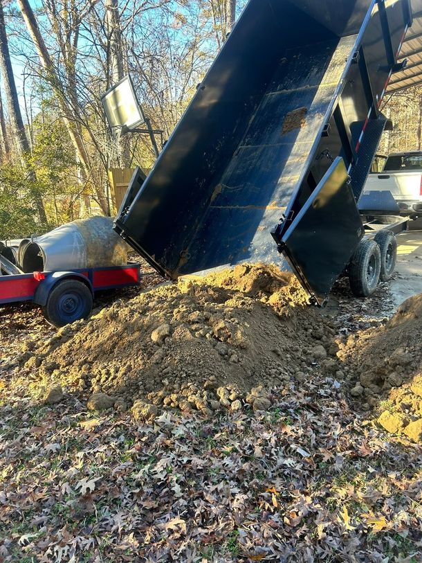 Dump truck unloading a large pile of dirt and leaves beside a red trailer in a wooded area
