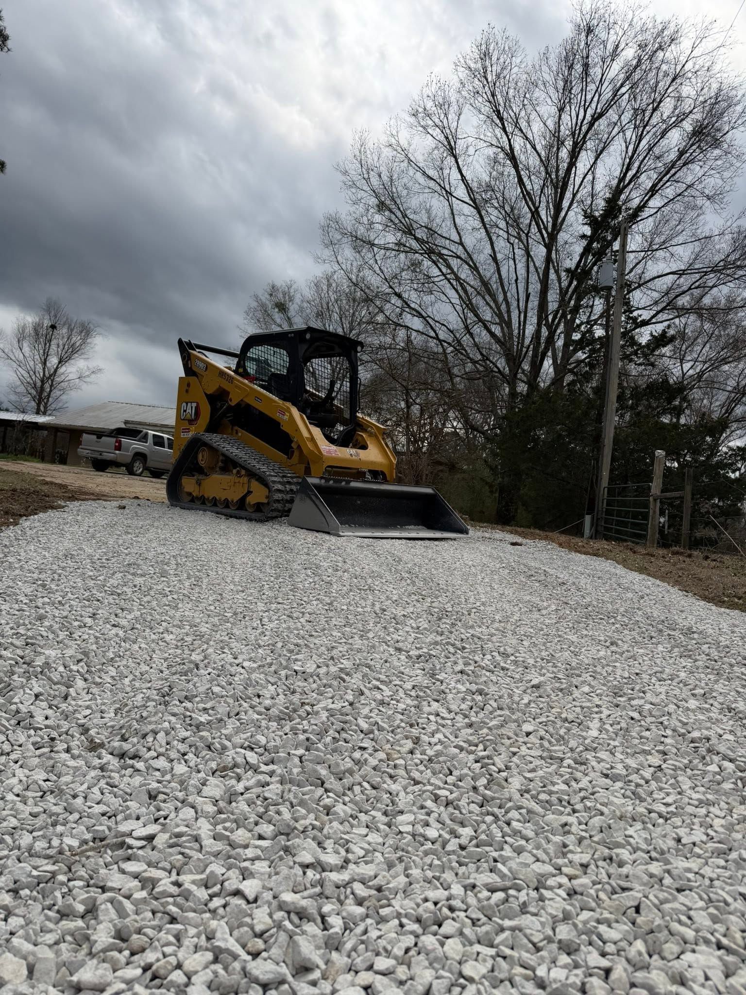 Yellow skid-steer loader on a gravel pile under cloudy skies, with trees in the background