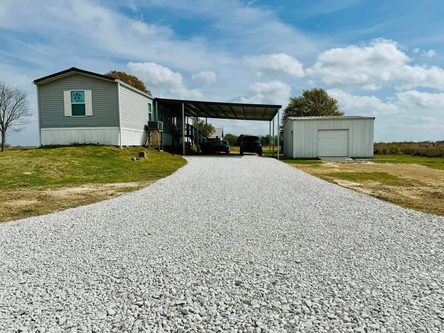 Gravel driveway leading to a small white house and garage under a cloudy sky.