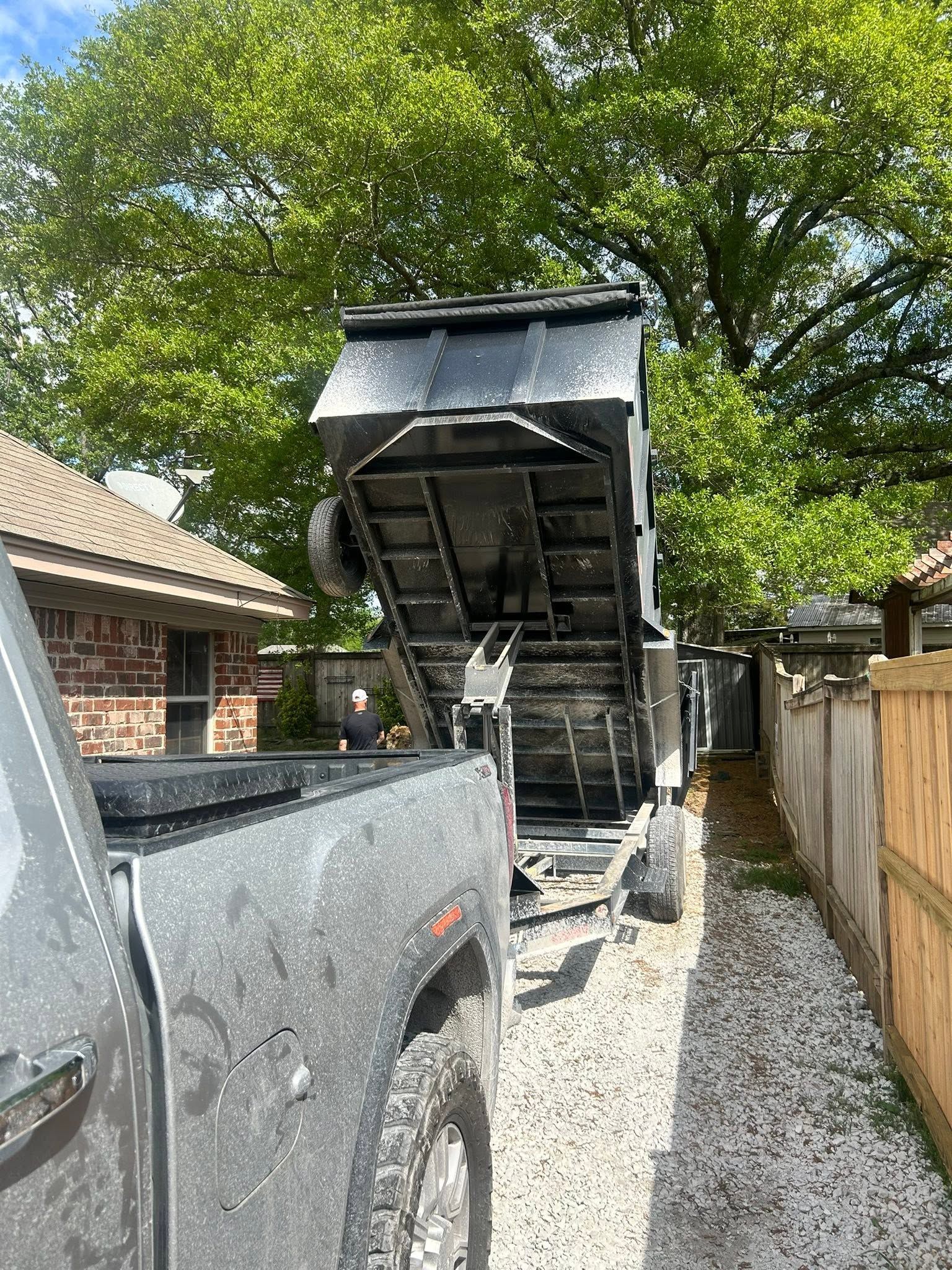 Truck dumping gravel into a narrow driveway beside a house, with a raised trailer and trees overhead