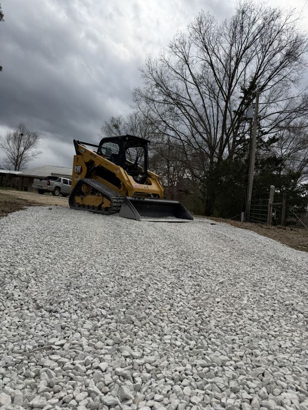 Yellow skid-steer on a large gravel mound under cloudy skies, with trees in the background