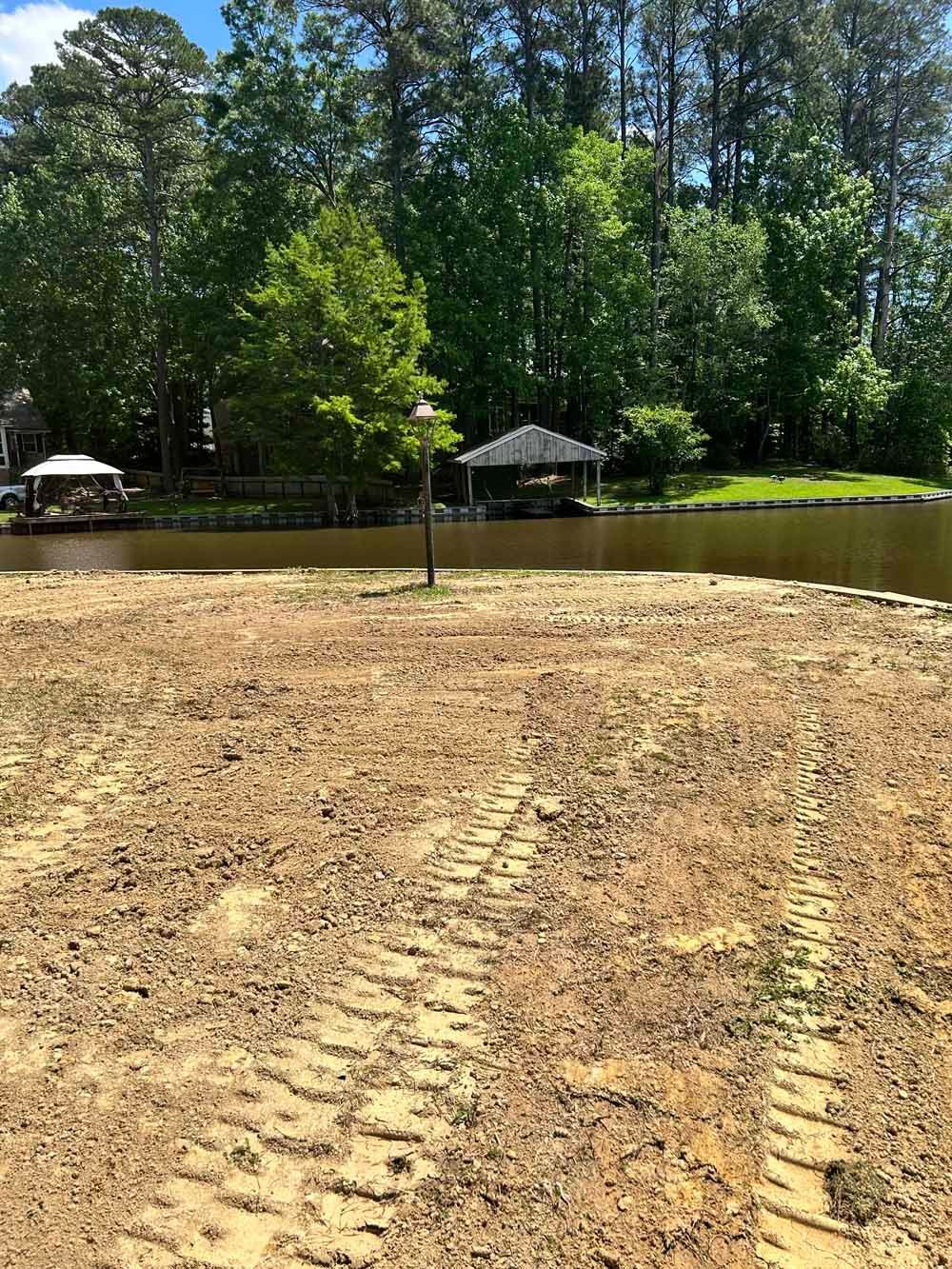 Dirt lot beside a pond with trees and a grassy bank under bright sun