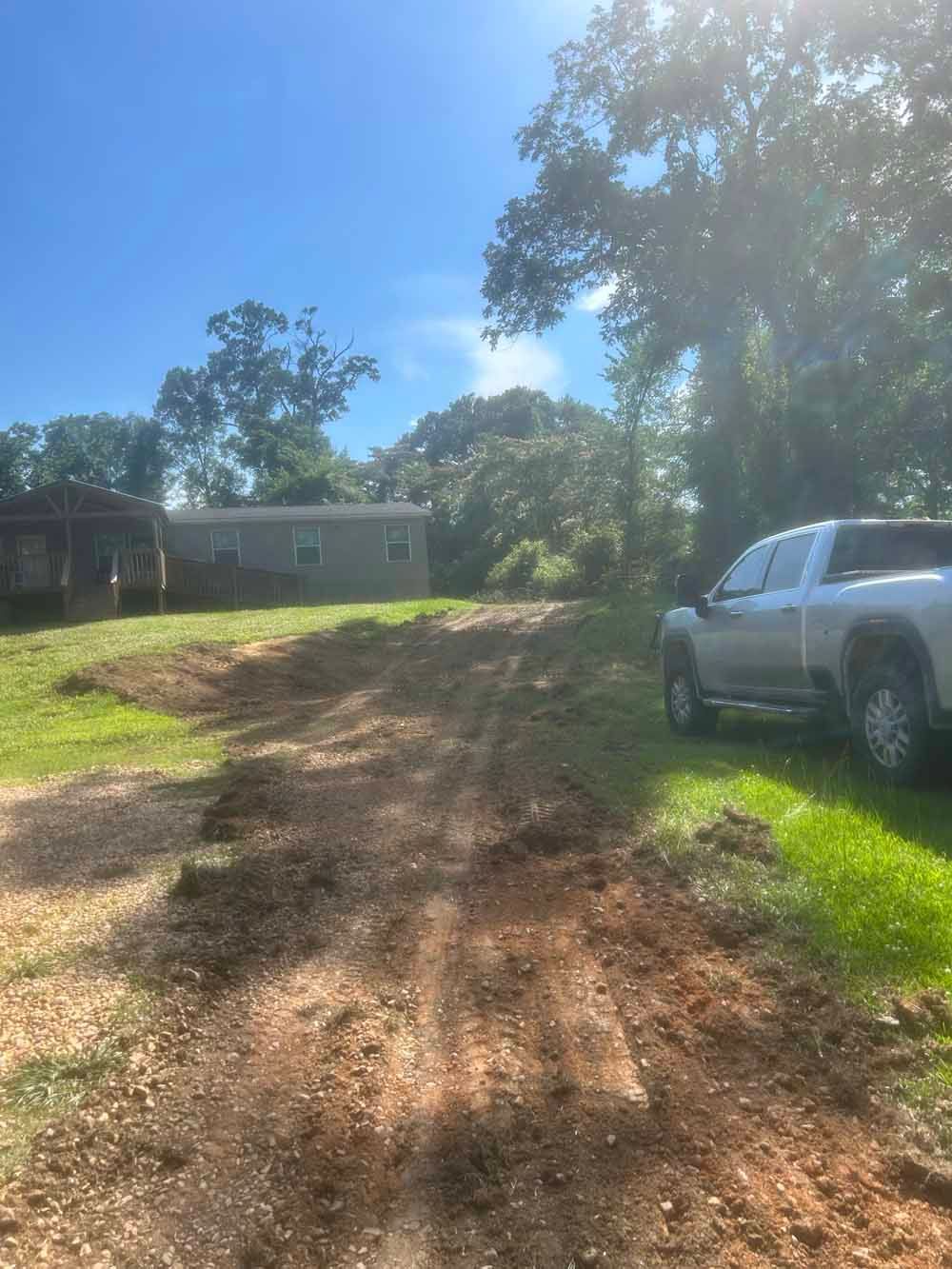 Dirt driveway leading to a house, with a silver SUV parked on the right and trees in bright sunlight.